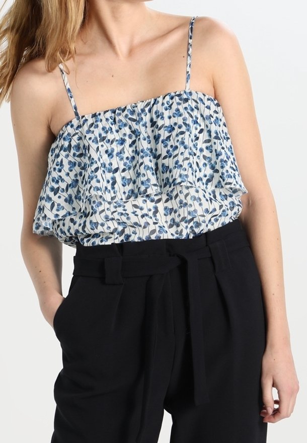 Femme portant un chemisier blanc sans manches avec un motif floral bleu et des volants, assorti à un pantalon noir taille haute avec une ceinture en tissu.