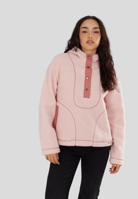 Vrouw in een lichtroze fleecepullover met een capuchon, knoopsluiting en lichtroze stiksels, gecombineerd met zwarte broek.