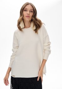 Kobieta z falującymi włosami do ramion nosi oversize'owy kremowy sweter, kolczyki z frędzlami i czarną koronkową spódnicę, pozując na białym tle.