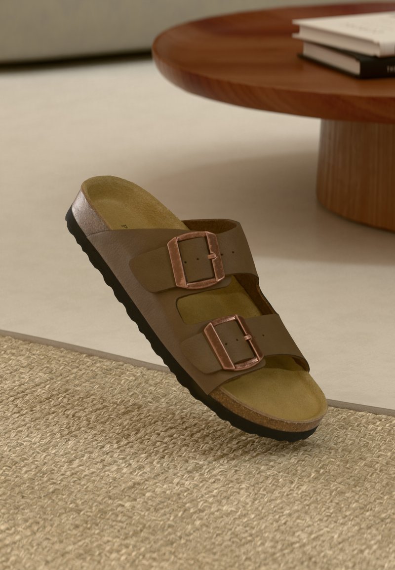 Bruna lädersandaler med två justerbara spännen, korkfotbädd och svart sula, utställda på en beige vävd matta nära ett träbord.