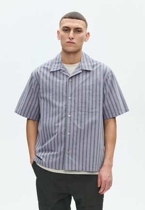 Joven con cabello corto que lleva una camisa gris de manga corta con botones y rayas negras verticales, de pie con una mano en el bolsillo.