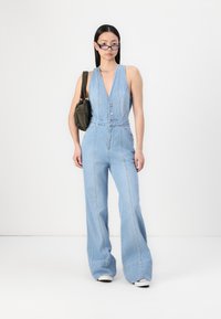 Une combinaison en denim bleu clair avec un décolleté en V profond, une fermeture à boutons sur le devant et des jambes larges. Associée à un sac en tissu vert olive et des lunettes.
