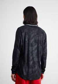 Camisa preta de manga longa com um padrão sutil de logótipos da Nike por toda a peça, com colarinho redondo e textura de tecido lisa e suave.