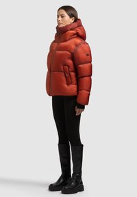 Czerwona puchowa kurtka z wysokim kołnierzem i kapturem, z zamkiem błyskawicznym i kieszenią po lewej stronie. Noszona z czarnymi dopasowanymi spodniami i wysokimi czarnymi botkami.