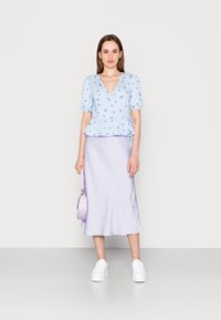 Top azul claro envolvente com estampa floral e mangas bufantes, combinado com uma saia midi lilás e ténis brancos de plataforma, segurando uma mala pastel.