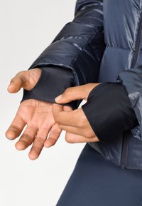 Puffer jacket featuring a navy exterior, zipper closure, and thumbhole cuffs. Smooth, glossy texture with quilted design. Close-up of hands adjusting cuffs.