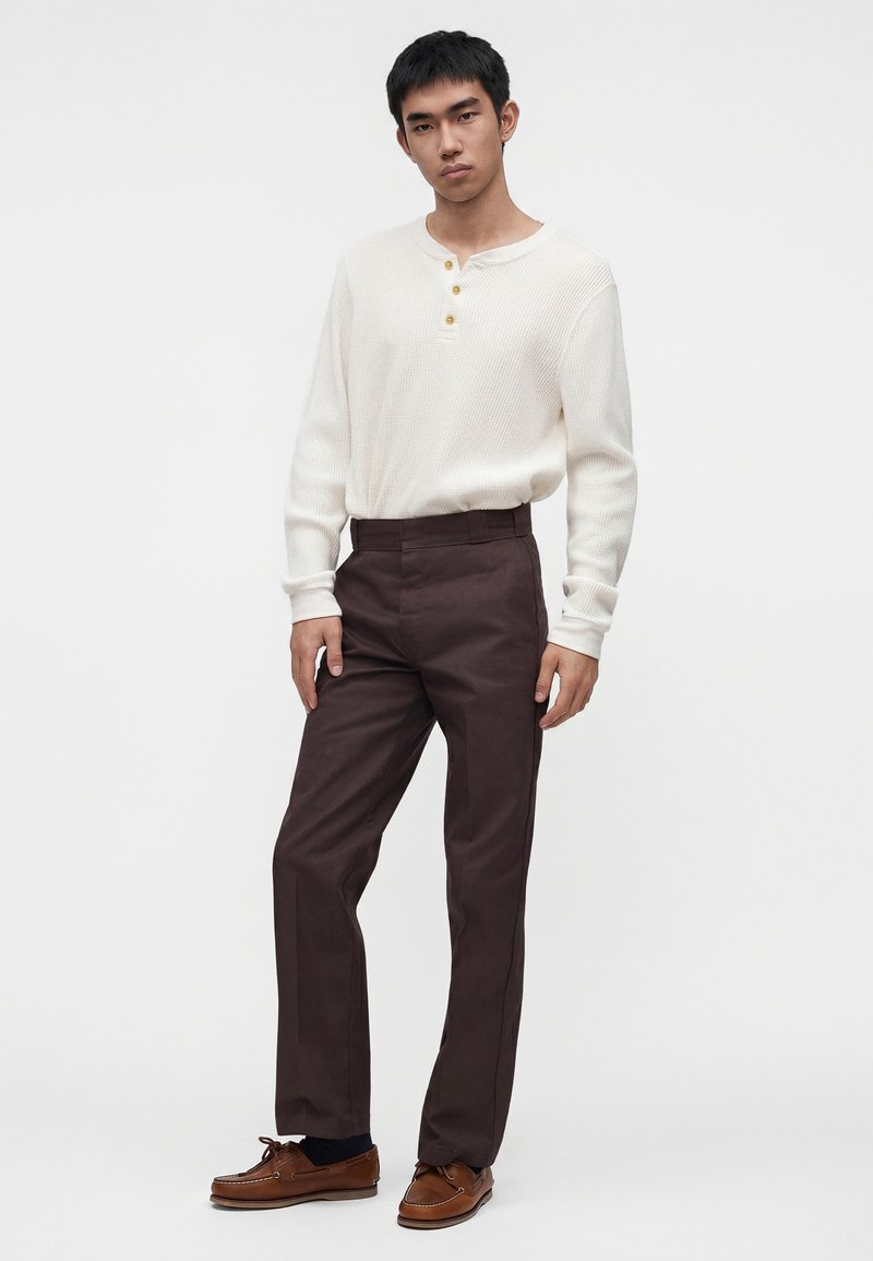 Giovane uomo in piedi, indossa una maglia henley bianca a maniche lunghe, pantaloni marrone scuro, calzini neri e scarpe da barca in pelle marrone.