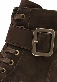 Brown suede ankle boot featuring a strap with a gold buckle, round toe, metallic eyelets, and brown lace. Textured surface detail visible.
