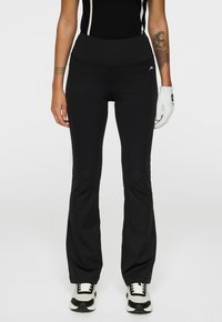 Persona che indossa pantaloni neri a zampa d'elefante, top nero senza maniche, guanto da golf bianco sulla mano destra e sneakers bianche e nere, in piedi su uno sfondo bianco.