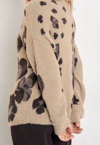 Pull beige en tissu doux et texturé avec un motif léopard noir. Il a des manches longues et une coupe décontractée, avec un ourlet arrondi.