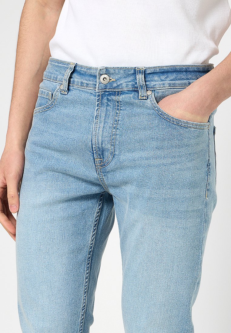 Persona con jeans de mezclilla azul claro y una camiseta sin mangas de rayas blancas, con una mano en el bolsillo delantero y la otra relajada a su lado.