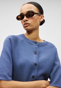 Μπλε πλεκτή ζακέτα με κοντά μανίκια, κούμπωμα με κουμπιά και στρογγυλή λαιμόκοψη. Συνδυασμένη με καστανά οβάλ γυαλιά ηλίου και απλά σκουλαρίκια.