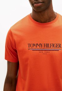 Camiseta de algodón naranja con cuello redondo. Presenta un logo en negro con el texto "TOMMY HILFIGER" y rayas debajo en azul, rojo y blanco.