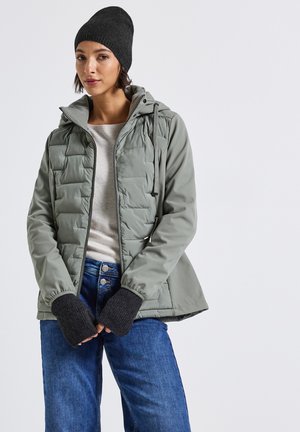 Giacca verde imbottita con cerniera, cappuccio regolabile e polsini elasticizzati. Indossata sopra una maglia leggera e abbinata a jeans blu. Berretto di maglia scuro.
