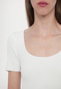 Valkoinen ribattu lyhythihainen paita, jossa on leveä suorakaiteen muotoinen kaula-aukko. Siinä on sileä pinta ja tiukka muotoilu, joka korostaa minimalistista tyyliä.
