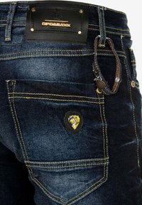 Jeans de mezclilla azul con un parche de cuero con el logotipo, costuras amarillas, un bolsillo pequeño y un emblema negro con acentos amarillos en la parte posterior.