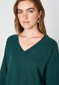 Maglione teal con scollo a V che presenta una vestibilità leggermente ampia e un tessuto a maglia testurizzato con maniche inserite. Dettaglio notevole è il collo pulito.