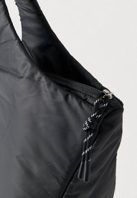 Sac en nylon noir avec un dessus zippé et une tirette de fermeture éclair en corde noire et blanche nouée, sur un fond clair uni.