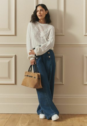 Woman wearing a white knit sweater and wide-leg blue jeans, holding a tan handbag, standing against a beige wall with panel molding.