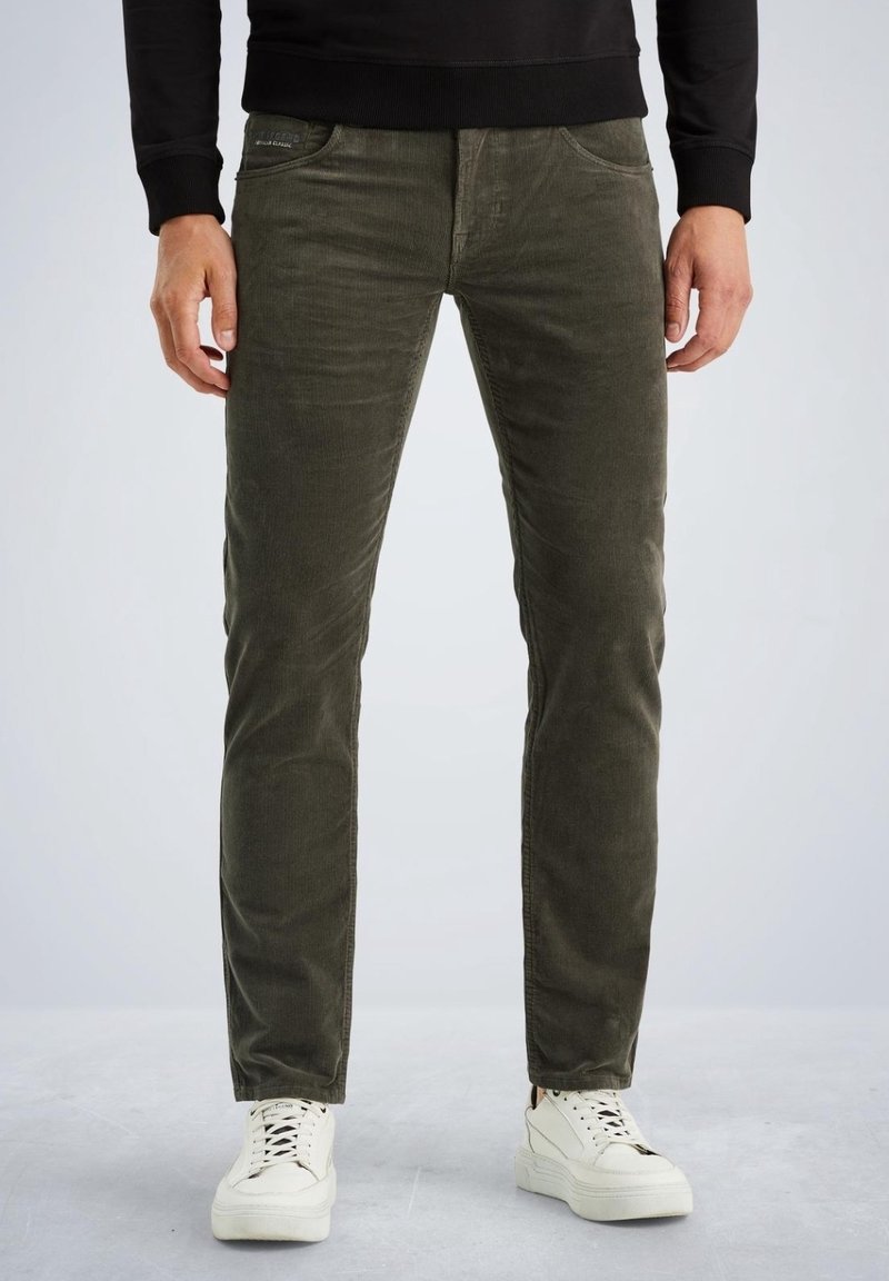 Homme portant un pantalon en velours côtelé vert olive, des baskets blanches et un t-shirt noir à manches longues, debout, les mains détendues le long du corps.