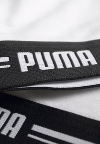 Černý tkaný pásek s bílým textem "PUMA", na kterém je vidět texturovaný povrch a plochý tvar. Pozadí je bílá tkanina.