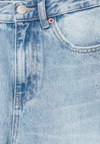 Jean en denim bleu clair en gros plan montrant la ceinture, les passants de ceinture, le bouton métallique, le rivet et la poche avant avec des détails de couture visibles.