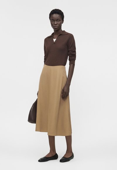 Γυναίκα που φοράει καφέ μπλούζα με μακριά μανίκια, μπέζ μίντι φούστα, μαύρα παπούτσια φλατ και κρατά μία σκούρα καφέ τσάντα, στέκεται μπροστά σε μονόχρωμο φόντο.