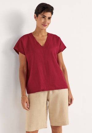 Kvinde står iført en rød V-hals kortærmet top og beige knælange shorts mod en ensfarvet lys baggrund.