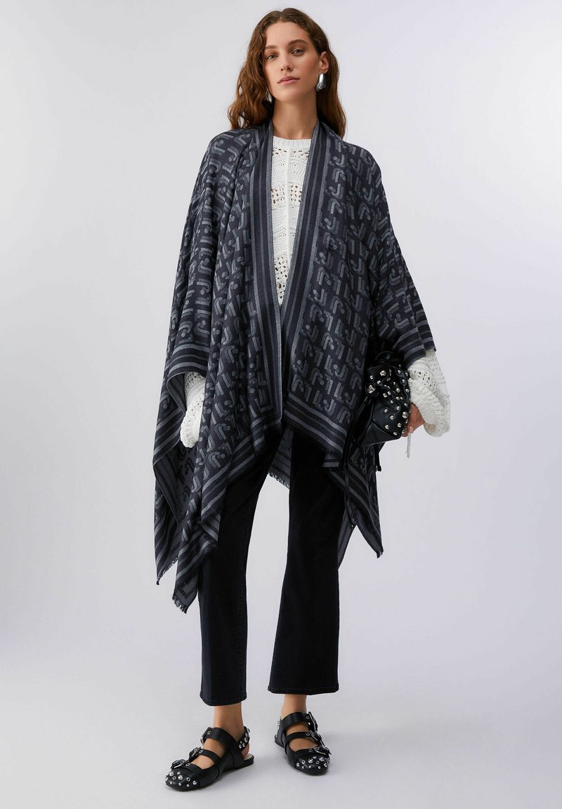 Nő fekete mintás sállal a fehér kötött pulóver fölött, fekete rövidnadrágban, szegecselt fekete szandálban, és egy fekete szegecselt táskát tartva.