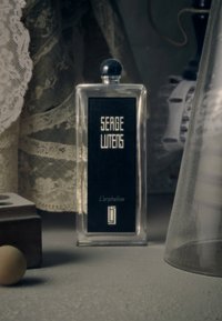 Bottiglia di profumo in vetro trasparente con un'etichetta nera che riporta "SERGE LUTENS" e "L'orpheline." Cappuccio rotondo nero. Ambientata su pizzo e vari oggetti.