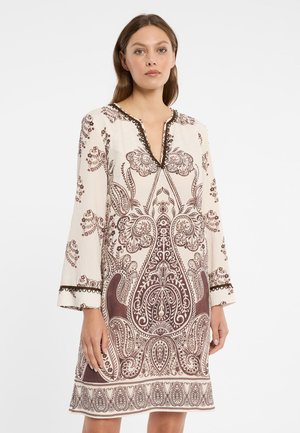 Frau trägt ein cremefarbenes, knielanges Kleid mit langen Ärmeln und einem aufwendigen braunen Paisley-Blumenmuster, stehend vor einem weißen Hintergrund.