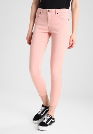Benendeel met lichtroze skinny jeans met rafelige zoom en zwart-witte lage sneakers, tegen een effen lichte achtergrond.