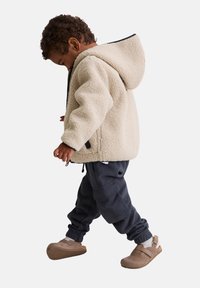 Roomkleurig fleecevest met capuchon, gecombineerd met donkerblauwe cargobroek en bruine suède schoenen. De outfit heeft contrasterende zwarte accenten.