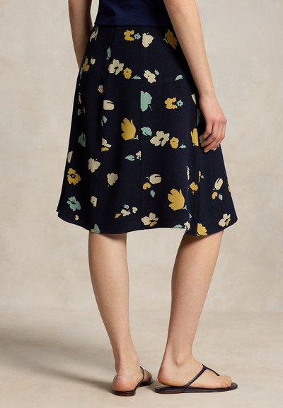 Femme portant une jupe bleu marine longueur genoux avec un imprimé floral jaune, beige et vert, associée à des sandales noires de type tong, vue de dos.