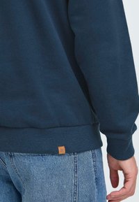 Persona che indossa un felpa blu scuro e jeans blu, mostrata dalla vita in giù, con la mano rilassata al lato.