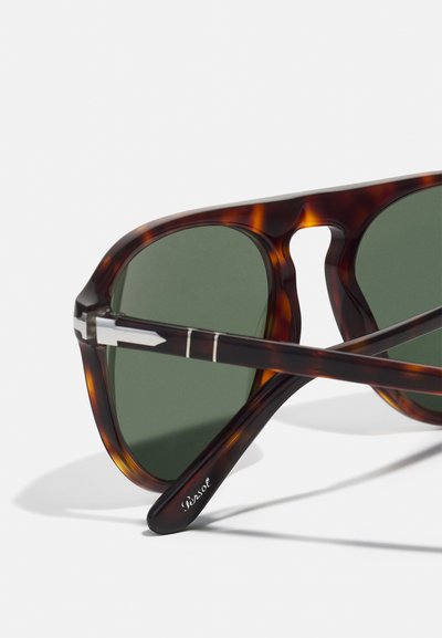 Sunglasses v tvare tortoiseshell s greenými šošovkami, ktoré majú geometrický dizajn, kovové akcenty na ráme a diskrétne logo na ramene.