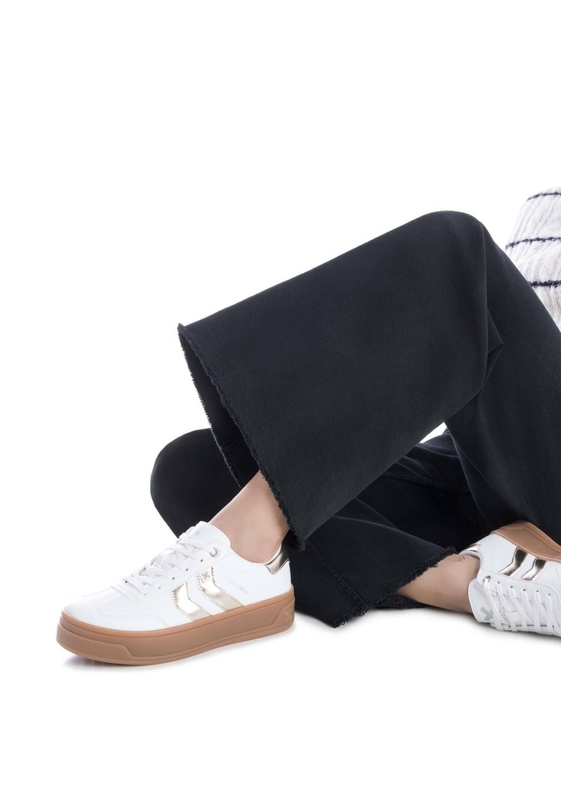 Zapatillas deportivas blancas con detalles dorados, suelas de goma texturizadas y cordones, combinadas con pantalones de pierna ancha negros con dobladillos deshilachados.