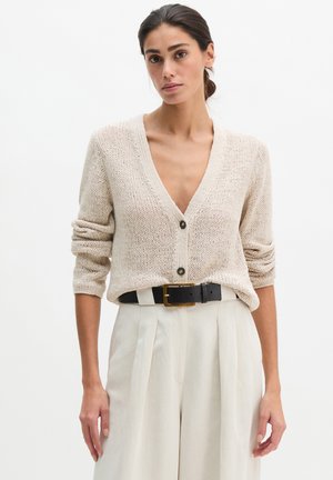 Frau mit zurückgebundenen dunklen Haaren, die eine beige Strickjacke mit Knöpfen und hoch taillierte cremefarbene Hosen mit schwarzem Gürtel trägt.