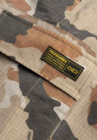 Tessuto mimetico con motivi beige, nero e marrone. Presenta un'etichetta del marchio "PROPAGANDA" in giallo e nero cucita sulla parte superiore sinistra.