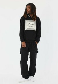 Sudadera negra con un parche de logo blanco y texto, combinada con pantalones cargo negros y zapatillas blancas. Presenta un corte relajado y un diseño sencillo.