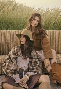 Deux femmes sont assises sur un canapé rayé multicolore. L'une porte un chapeau beige et un pull brun à motifs. L'autre a une chemise brune et des bottes jusqu'aux genoux.