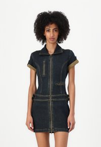 Denimdress med kortermede ermer, en frontglidelås og to lommer. Har et mørkeblått stoff med kontrasterende gule sømmer og mansjettdetaljer.