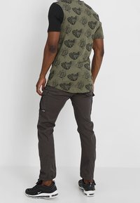 Olivgrünes kurzärmliges Hemd mit schwarzen Ärmeln und Blättermuster, kombiniert mit dunkelgrauen Cargo-Hosen und schwarzen Sportschuhen mit weißen Akzenten.