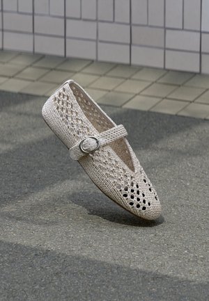 Zapato beige tejido con diseño perforado, una abertura en forma de V y una correa decorativa con una hebilla circular plateada. Suela plana.