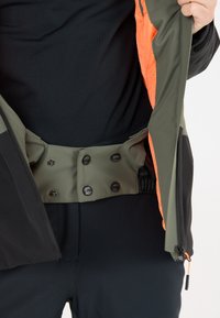 Sort og grøn jakke med en orange inderside, der har tryknapper og en elastisk talje. Materialet ser glat og letvægtigt ud.