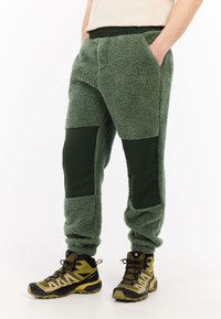 Groene fleecebroek met donkergroene nylon patches op de knieën, een elastische tailleband en zijzakken. Gecombineerd met zwarte en gele wandelschoenen.