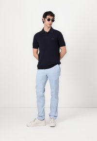 Polo shirt en tricot marine à manches courtes, pantalon bleu clair et baskets blanches. Modèle portant des lunettes de soleil noires, se tenant devant un fond neutre.