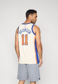 Basketballtrikot in Cremefarbe mit blauen und orangefarbenen Akzenten, auf der Rückseite mit "BRUNSON" und der Nummer "11" versehen, aus leichtem Stoff und ärmellosem Design.
