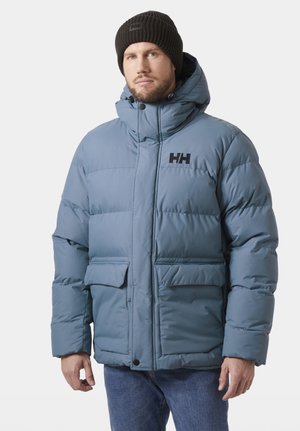 Man draagt een lichtblauwe gevoerde winterjas met capuchon en een zwarte muts, staand tegen een effen lichte achtergrond.