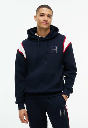 Homme portant un sweat à capuche bleu marine et un pantalon assorti avec des bandes rouges et blanches et des écussons au logo "Hilfiger" sur la poitrine et la jambe.